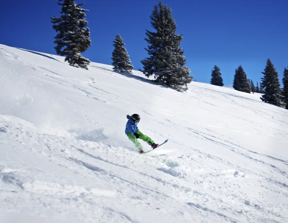 A snowboarder in a blue jacket kicking up powder on a bluebird day among snow-covered evergreens at Vail