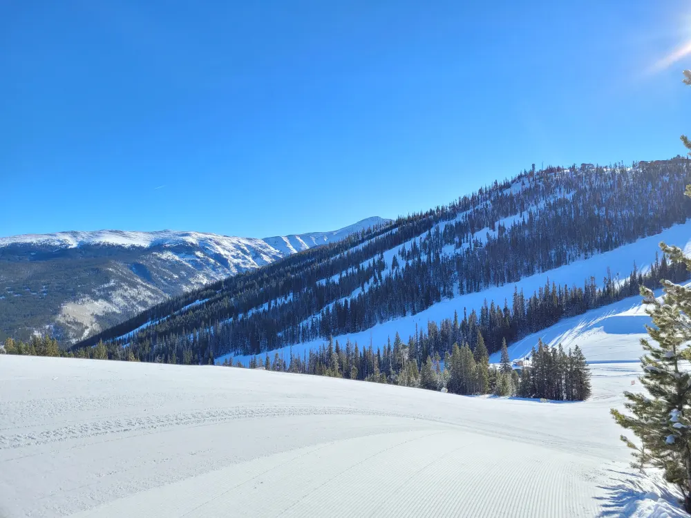 A freshly groomed corduroy run at Winter Park with treed slopes and the Continental Divide under a clear sky